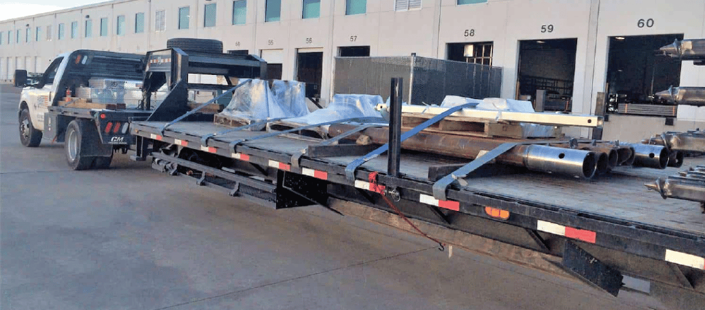 In a warehouse parking lot a pick up truck is attached to a gooseneck trailer hauling metal pipes and other material