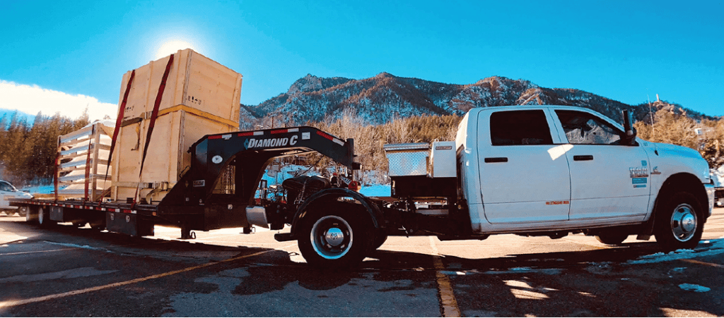 In a parking lot with mountains in the background, a pick up truck is attached to a gooseneck trailer loaded with wooden crates.