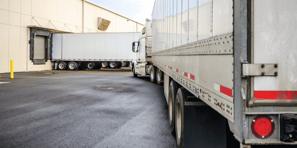 A white semitruck pulls into a loading dock next to another white truck