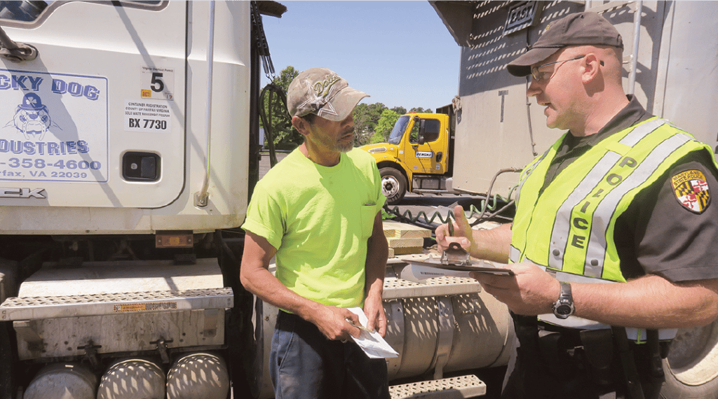 in front of a semi truck a truck driver in neon yellow speaks to a maryland police officer who is holding a clipboard